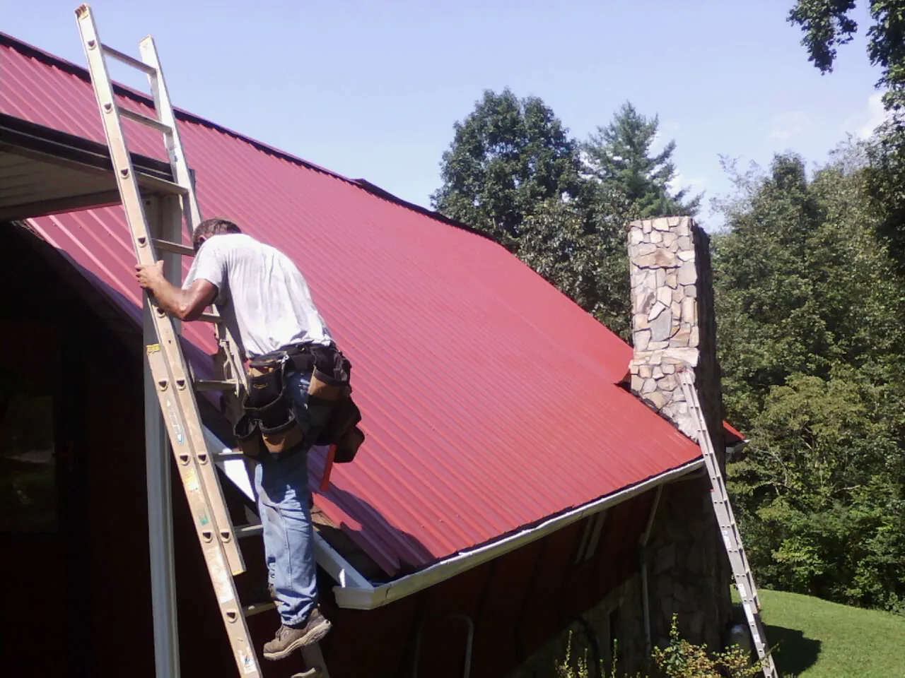 Roofer on a ladder beside a red metal roof and stone chimney — Soffit & Fascia Repair by Roofing Brothers in Glen
